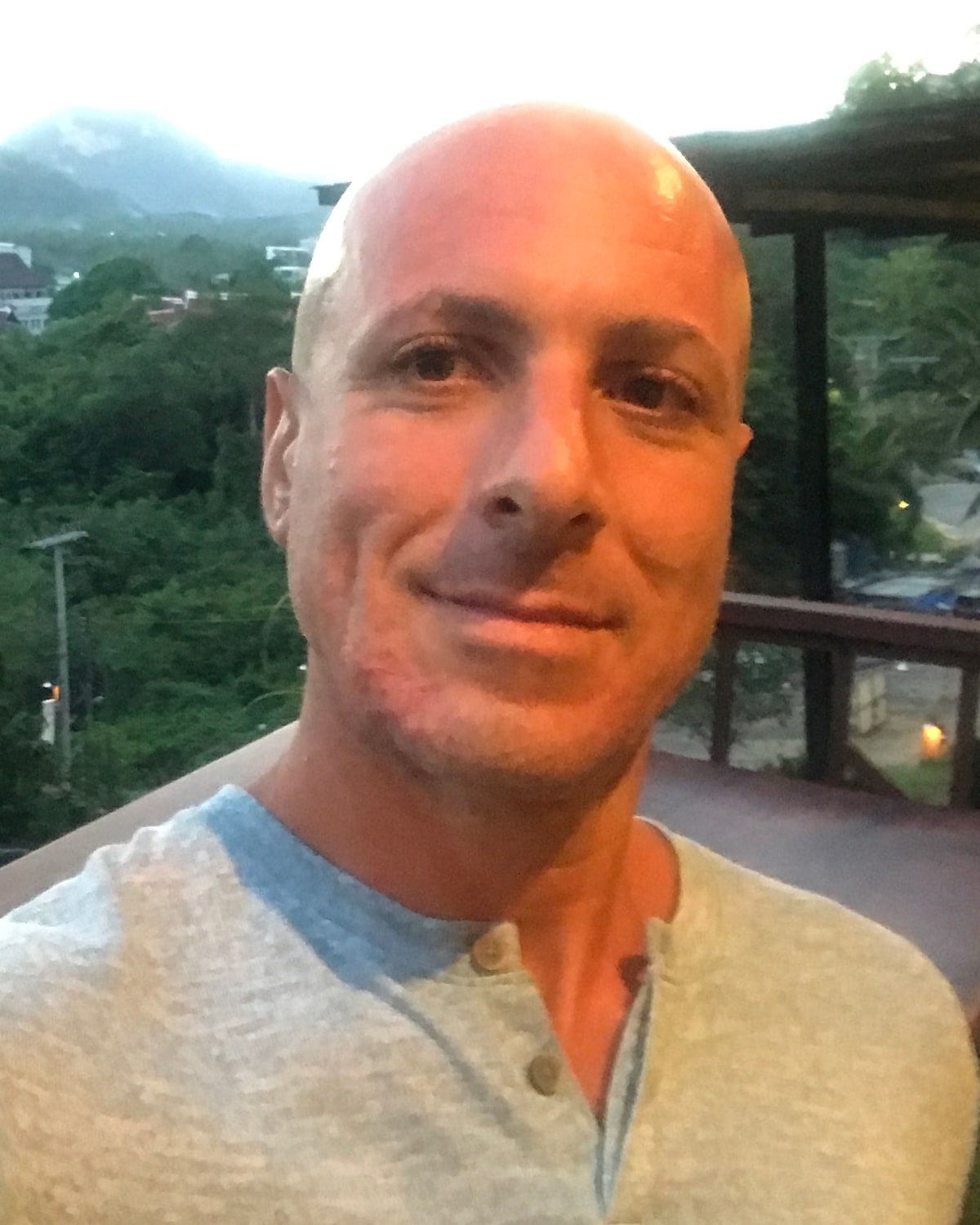Gabe, founder of The Quiet Scribe, smiling mid-thought on a hillside cafe terrace in Khanom, Thailand, in warm evening light with the town and mountains behind him.